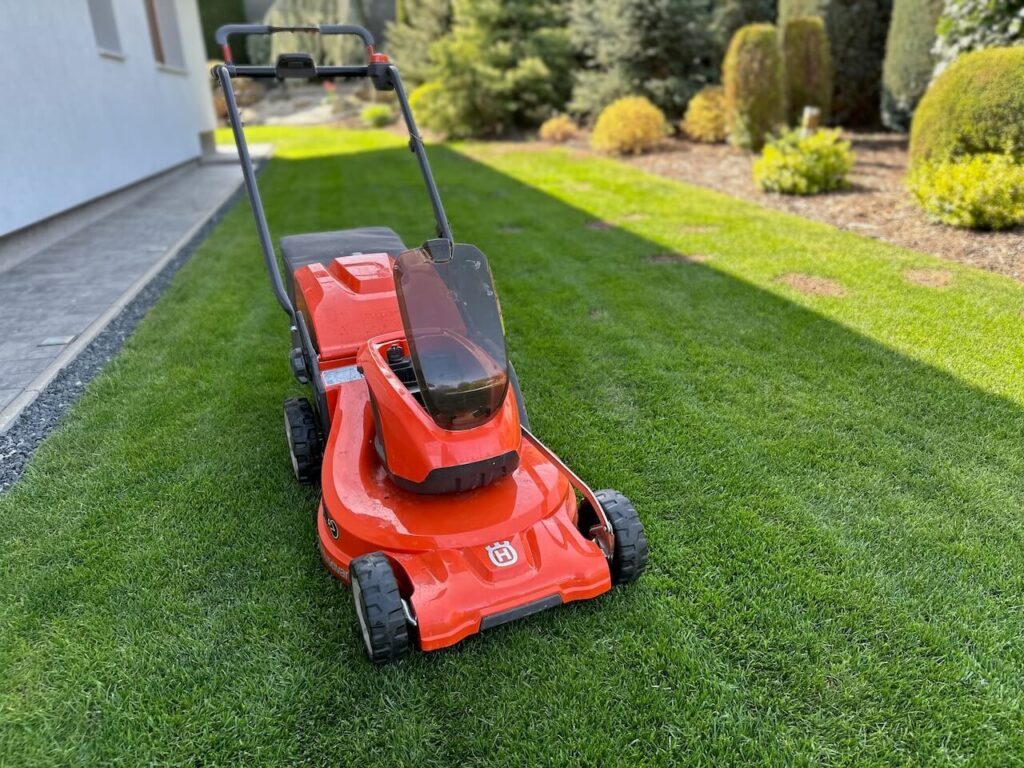 Sekačka na trávu oranžové barvy Husqvarna stojí na upraveném trávníku s výhledem na zahradu s pečlivě zastřiženými keři a stromy v pozadí.