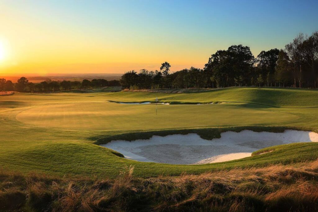 Západ slunce nad golfovým hřištěm s jemně zvlněnými fairwayemi, písečnými bunkry a zelenými plochami, v pozadí rozsáhlá krajina.