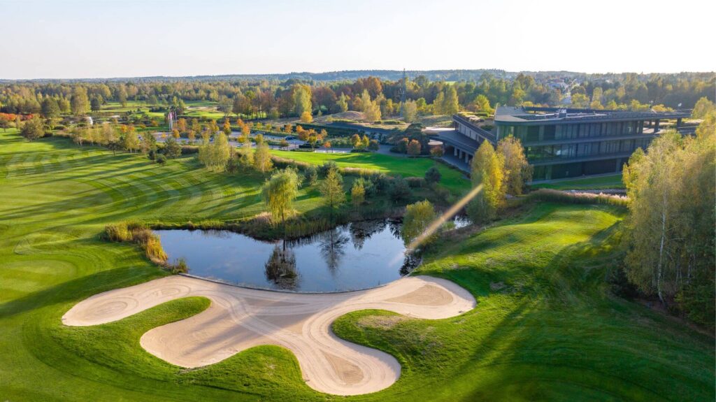 Aeriální pohled na golfový klub Rosa v Polsku, s jezerem, písečnými překážkami, rozlehlými zelenými fairways a moderním klubovým domem obklopeným stromy.