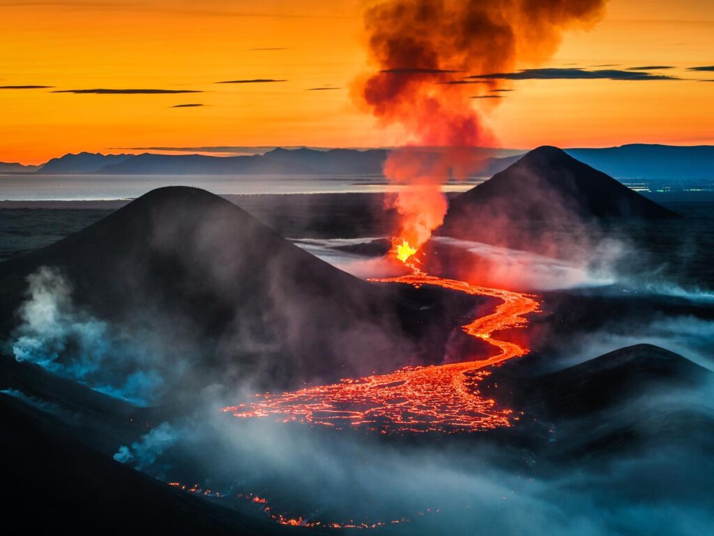 Erupce sopky při západu slunce, s tokem lávy svítícím v tmavém krajinném reliéfu a sloupy dýmu stoupajícími do barevně rozžhaveného nebe.