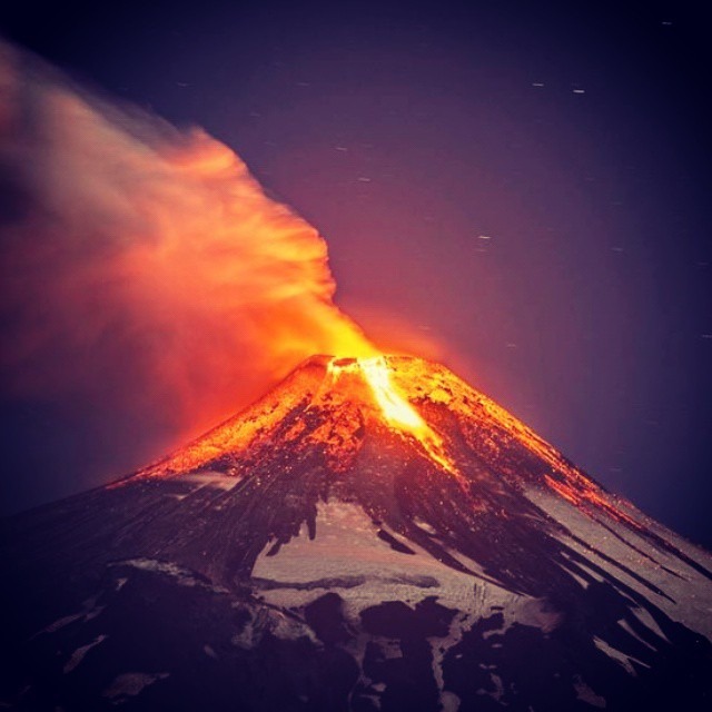 Noční fotografie erupce sopky Villarrica se světlou lávou stékající po svazích a oranžovým dýmem v kontrastu s tmavou oblohou.