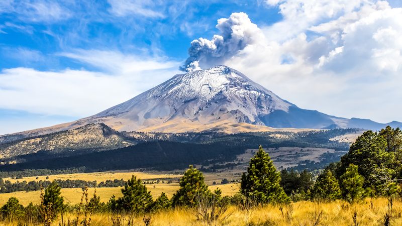 Sopka Popocatépetl v Mexiku s kouřem vystupujícím z kráteru, zasazená do idylické krajiny s modrým nebem a zasněženým vrcholem.