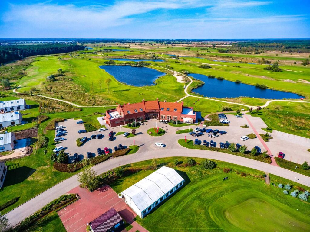 Pohled shora na golfový klub Sobienie Królewskie, s výhledem na zelené hřiště, jezera a klubovnu.
