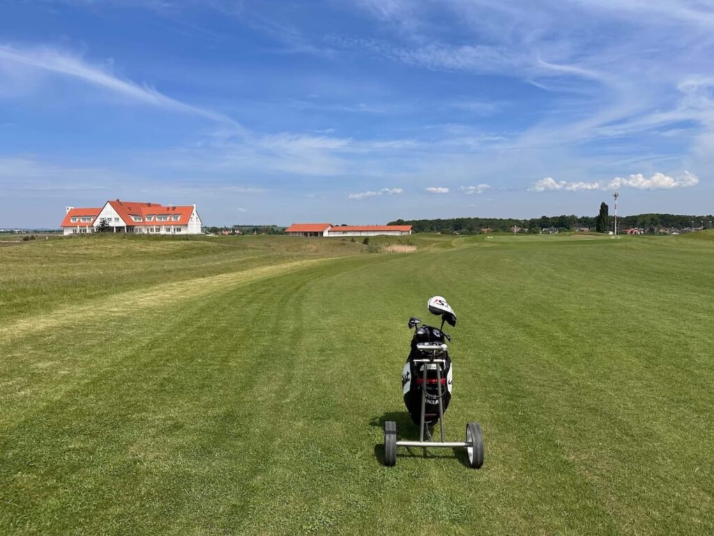 Golfový vozík Titleist stojí na fairwayi šesté jamky s výhledem na golfový klub a rozlehlé golfové hřiště ve Vinorě, Praha, pod jasně modrou oblohou.
