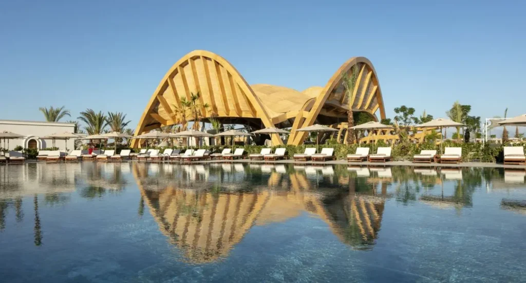 Luxusní hotelový resort v Beleku, Turecko, s moderní architekturou a velkým venkovním bazénem obklopeným lehátky a slunečníky. Hotelový design zahrnuje unikátní dřevěné struktury a bohatou zeleň, které vytvářejí atmosféru tropického ráje.