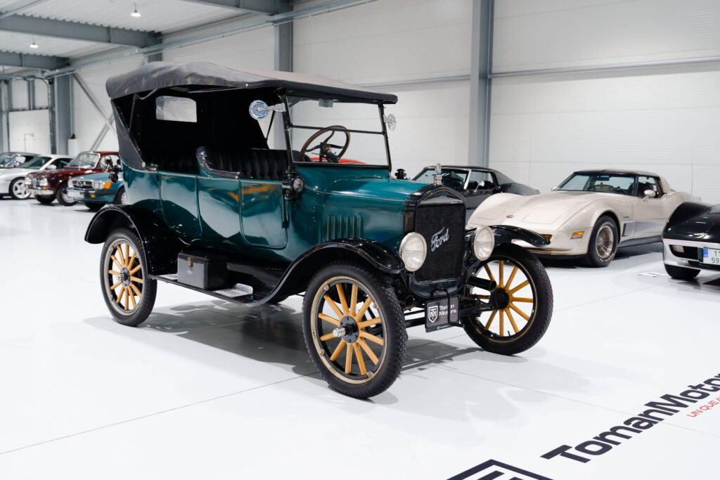 Historický Ford Model T Roadster z roku 1925 v elegantní zelené barvě, vystavený v moderní automobilové hale mezi dalšími klasickými vozy. Tento ikonický vůz má dřevěná kola a otevřenou karoserii, což ilustruje revoluční design a technologii své doby.