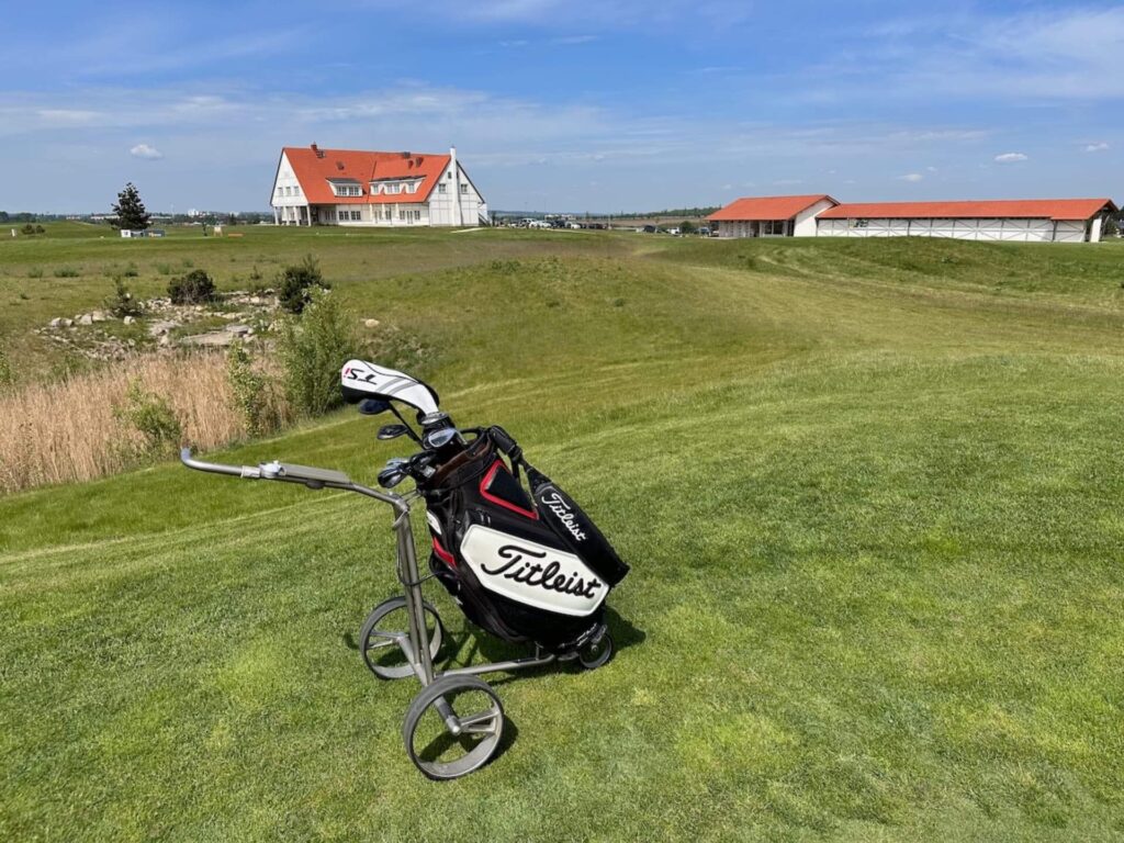 Golfový vozík značky Titleist stojící na zeleném fairwayi s výhledem na golfový klub a otevřenou krajinu v Golf City Praha Vinor.