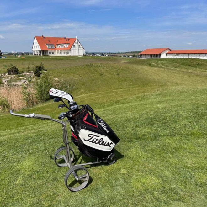 Golfový vozík značky Titleist stojící na zeleném fairwayi s výhledem na golfový klub a otevřenou krajinu v Golf City Praha Vinor.