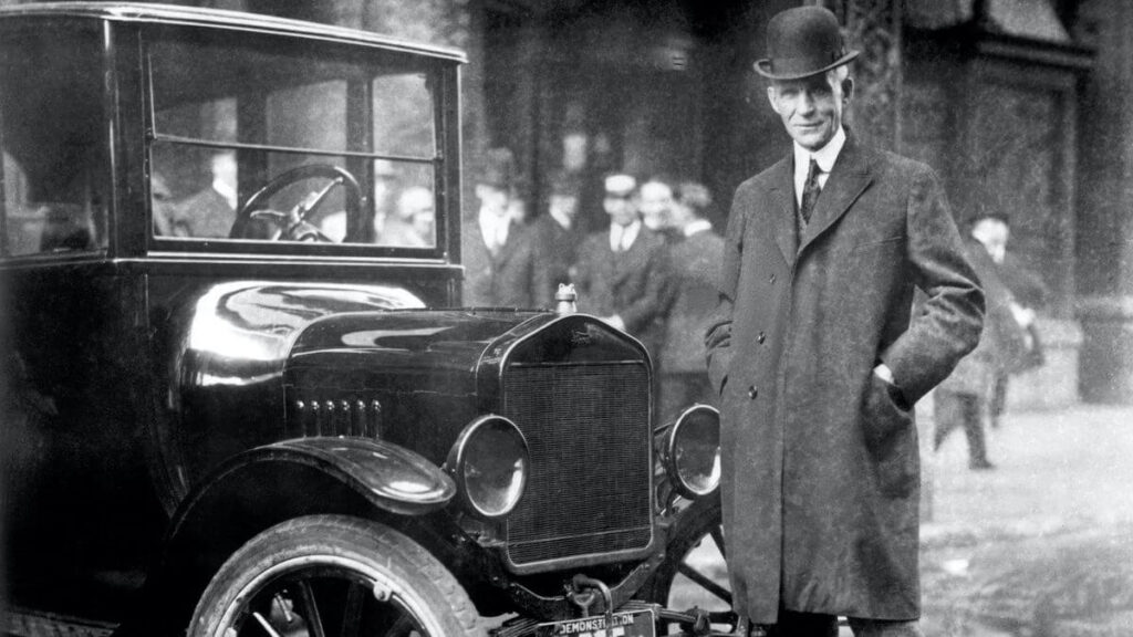 Henry Ford stojí vedle automobilu Model T, který revolučním způsobem změnil automobilový průmysl. Ford je oblečen v elegantním kabátě a klobouku, s úsměvem na tváři. Fotografie zachycuje historický okamžik v ulicích plných lidí, pravděpodobně z počátku 20. století.