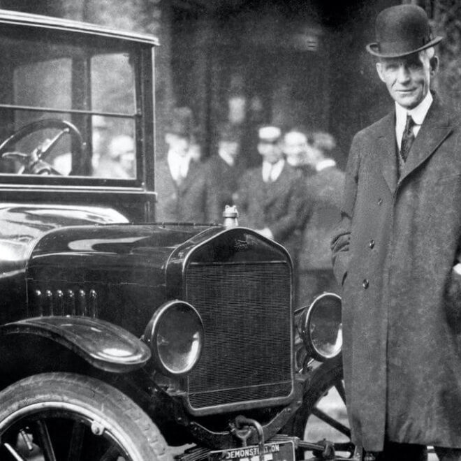 Henry Ford stojí vedle automobilu Model T, který revolučním způsobem změnil automobilový průmysl. Ford je oblečen v elegantním kabátě a klobouku, s úsměvem na tváři. Fotografie zachycuje historický okamžik v ulicích plných lidí, pravděpodobně z počátku 20. století.