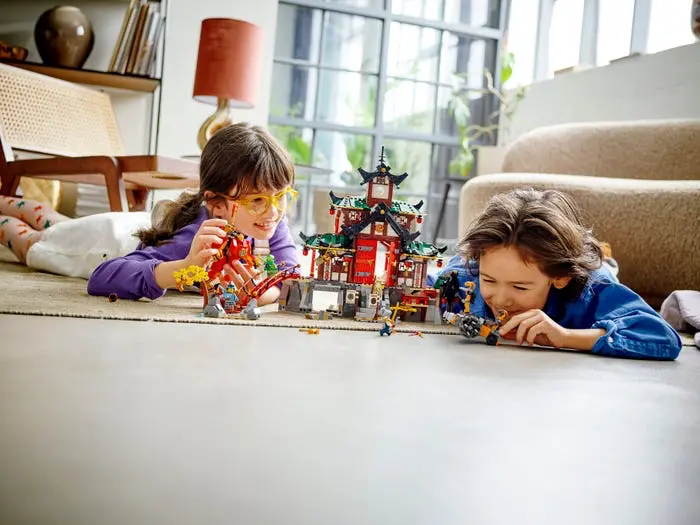 Dvě děti si hrají s Lego stavebnicí na podlaze obývacího pokoje. Dívka v brýlích a chlapec se zaujetím skládají a hrají si se stavebnicí, která připomíná asijský chrám.