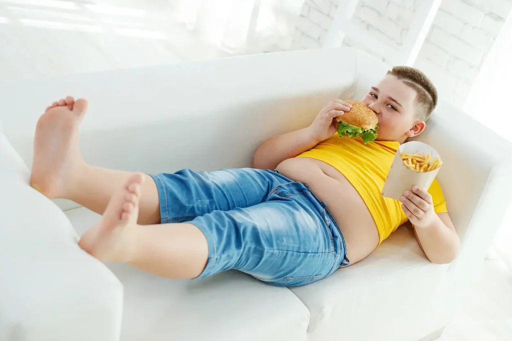 Chlapec ležící na pohovce, jí hamburger a hranolky, oblečený do žlutého trička a džínových šortek. Tento obrázek ilustruje problém dětské obezity a nezdravého životního stylu.