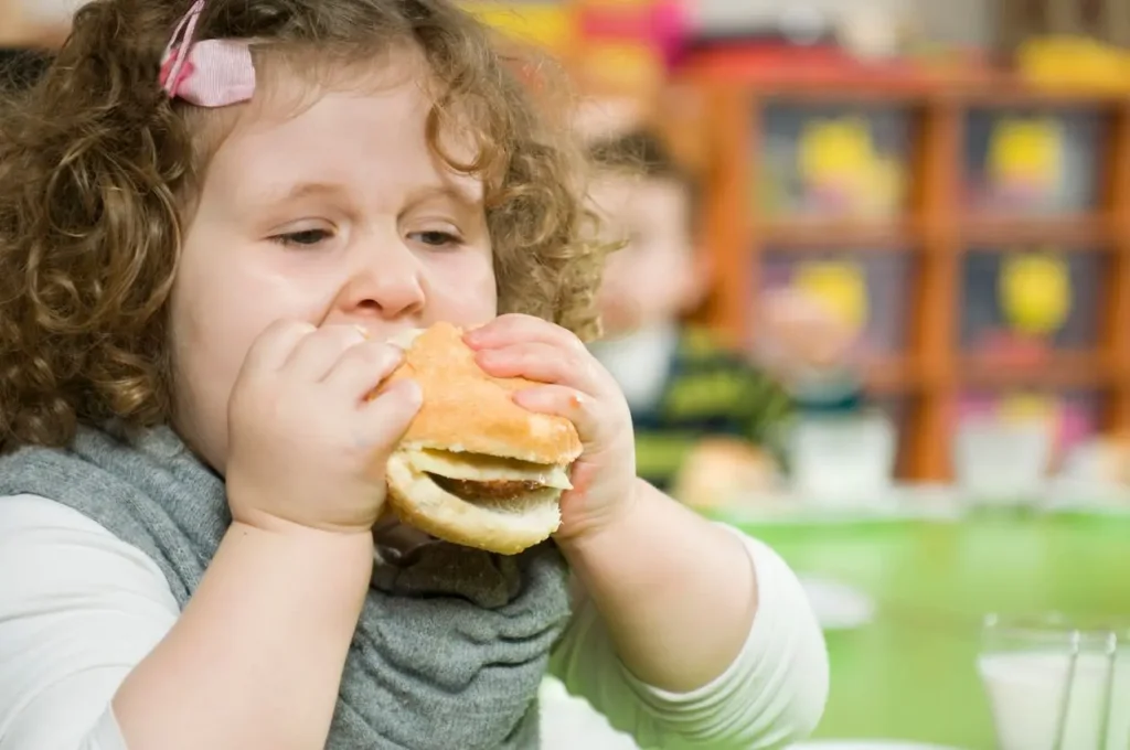 Holčička s kudrnatými vlasy a růžovou sponkou, která jí hamburger. Obrázek ilustruje problematiku dětské obezity a nezdravých stravovacích návyků.