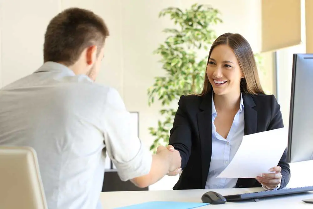 Žena podává ruku muži během pracovního pohovoru, usmívá se a drží papíry s poznámkami. Na pozadí je zelená rostlina a světlé prostředí kanceláře.