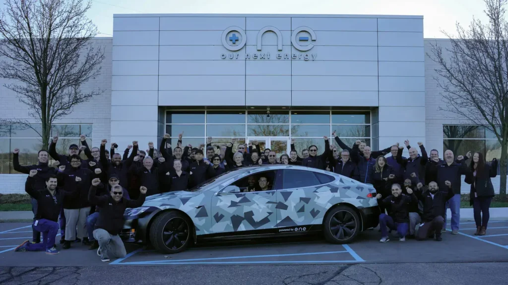 Skupinová fotografie týmu společnosti Our Next Energy (ONE) před jejich sídlem, spolu s elektromobilem Tesla, který je vybaven jejich revoluční baterií Gemini, schopnou dojezdu 965 km na jedno nabití.