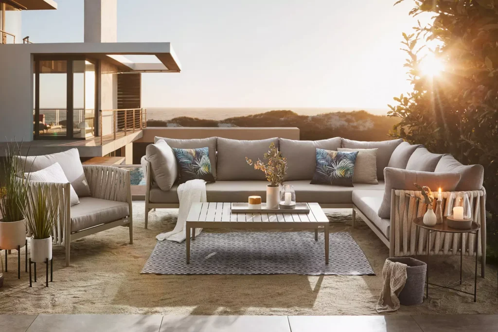 Moderní zahradní posezení s pohodlnou rohovou sedačkou a křesly v elegantním designu, situované na terase s výhledem na moře při západu slunce. Celý prostor je doplněn dekorativními polštáři, koberci a osvětlením, vytvářející útulnou atmosféru pro relaxaci.