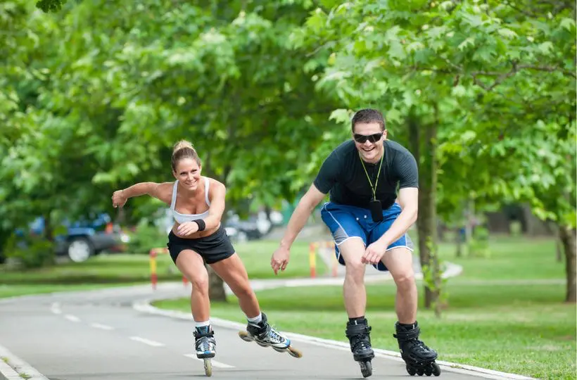 Dva lidé bruslí na kolečkových bruslích po cyklostezce v parku. Usmívají se a užívají si venkovní aktivitu za slunečného dne.