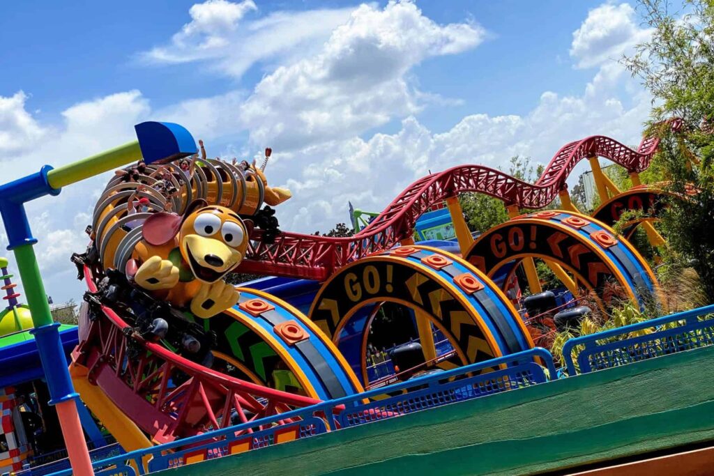 Barevná horská dráha ve tvaru hračky z filmu Toy Story se strmými zatáčkami a veselým designem. Horská dráha nese podobu roztažitelného psa a projíždí kolem nápisů "GO!". Na pozadí je modrá obloha s bílými mraky a okolní vegetace.