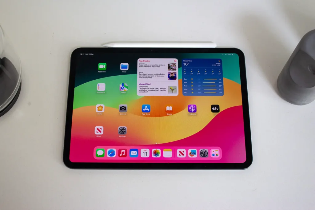 Apple iPad Pro 11 2024 s OLED displejem na bílém stole, zobrazená hlavní obrazovka s aplikacemi a widgety, nad tabletem leží Apple Pencil.