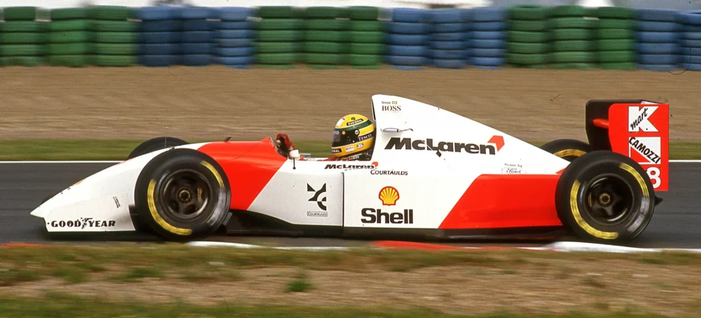 Ayrton Senna řídící svůj McLaren-Honda ve Formuli 1 během závodu, zachycený na okruhu, s jeho ikonickou žlutou helmou a červenobílým vozem.