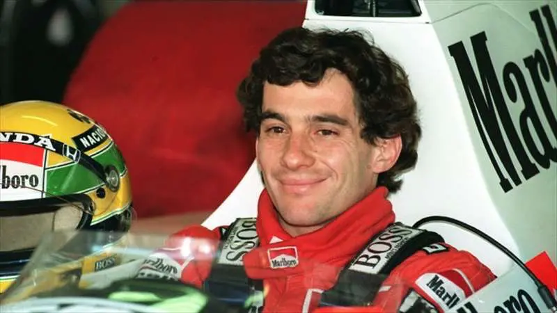 Ayrton Senna, legendární automobilový závodník Formule 1, sedící ve svém závodním voze, s úsměvem na tváři, vedle své ikonické závodní helmy.