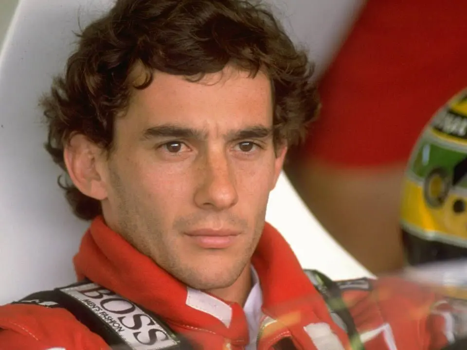 Ayrton Senna, slavný závodník Formule 1, soustředěný před závodem, v závodním obleku s jeho ikonickou žlutou helmou v pozadí.