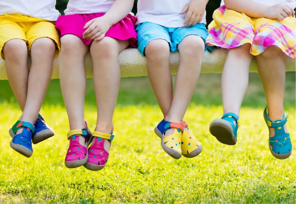Děti sedící na lavičce s nohama ve vzduchu, nosící barevné sandály. Oděny jsou v různě barevném oblečení, od žlutých kraťasů po růžové sukýnky, s trávou v pozadí.