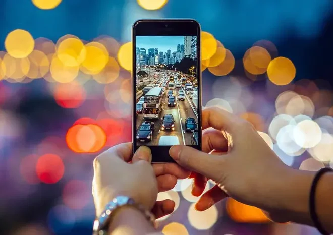 Osoba držící mobilní telefon s foťákem, fotografující večerní město s dopravní zácpou a rozmazanými světly v pozadí.