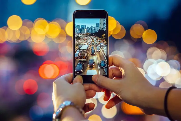 Osoba držící mobilní telefon s foťákem, fotografující večerní město s dopravní zácpou a rozmazanými světly v pozadí.