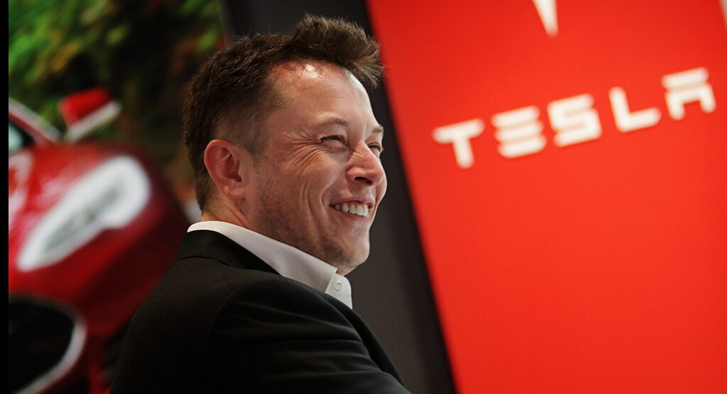 Elon Musk s úsměvem stojí před logem společnosti Tesla a červeným elektromobilem v pozadí.