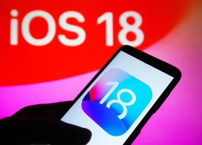 Obrázek ukazuje ruku držící iPhone s ikonou iOS 18 na obrazovce, v pozadí je vidět nápis "iOS 18".