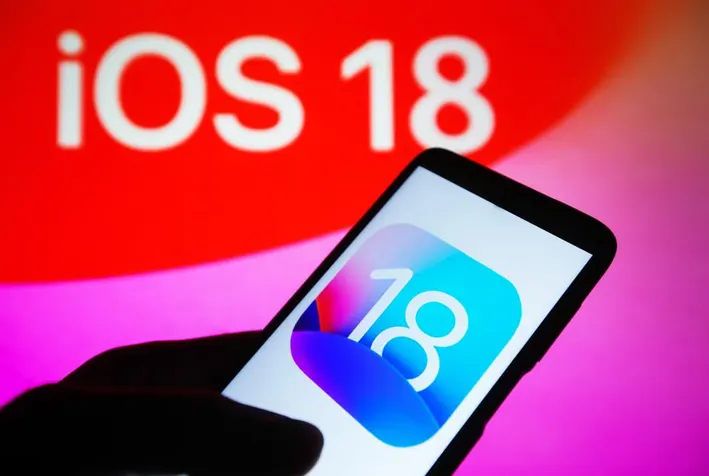 Obrázek ukazuje ruku držící iPhone s ikonou iOS 18 na obrazovce, v pozadí je vidět nápis "iOS 18".