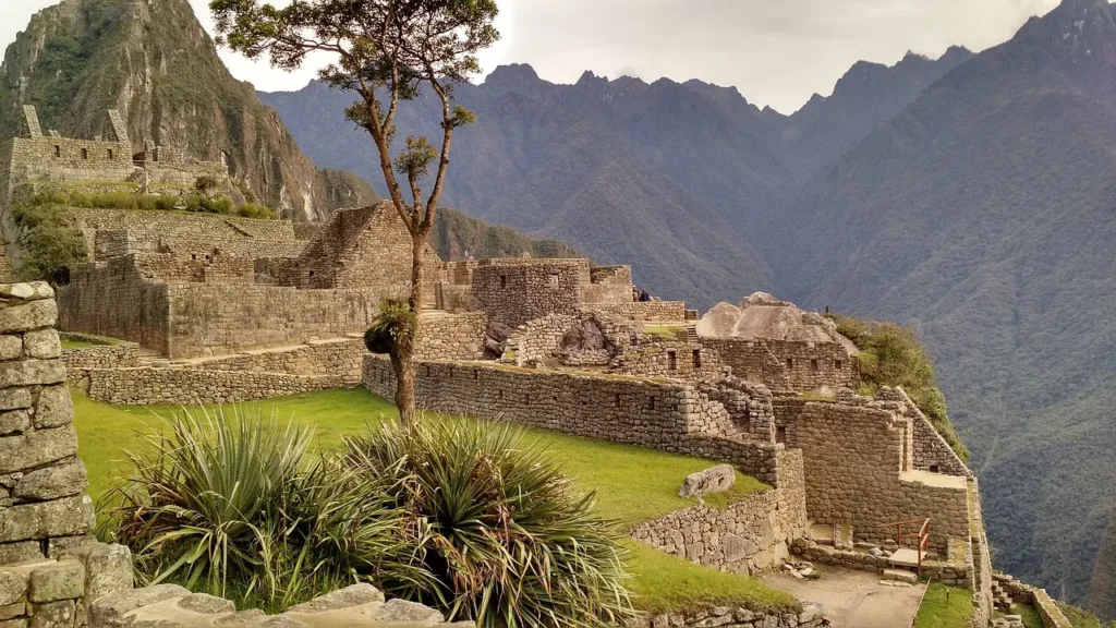 Detailní pohled na ruiny Machu Picchu v Peru, s kamennými strukturami, stromy a horami v pozadí, ukazující mistrovskou inckou architekturu.