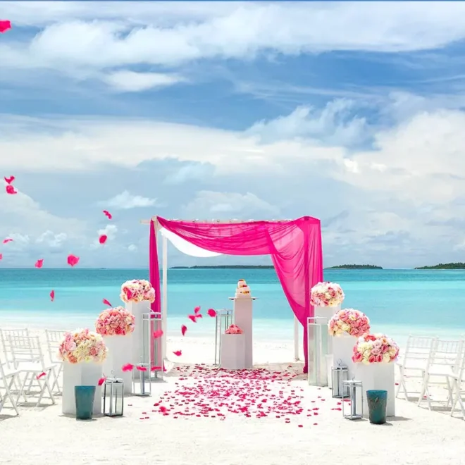 Romantická svatební scéna na pláži Malediv s bílým pískem, tyrkysovým mořem a růžovou dekorací. Červené růžové lístky jsou rozesety po pláži a víří ve vzduchu, zatímco bílé židle jsou uspořádány před obřadním altánem s růžovým závěsem.