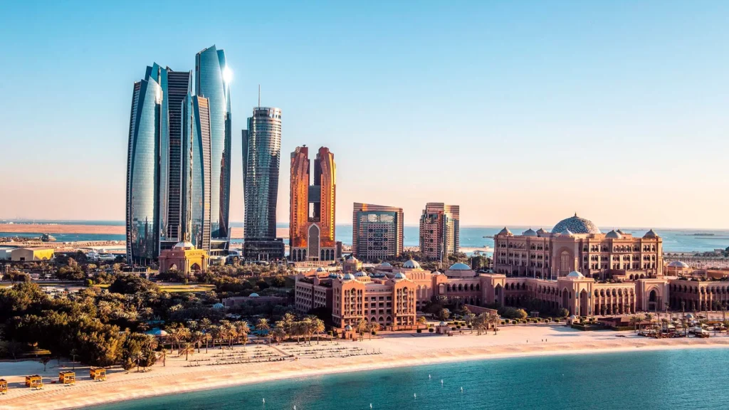 Moderní panorama Abu Dhabi zahrnující ikonické mrakodrapy Etihad Towers a luxusní hotel Emirates Palace, ležící přímo u pláže s modrým mořem v popředí.