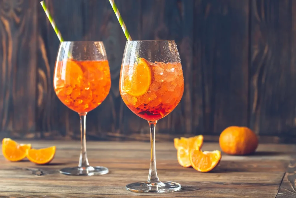 Dvě sklenice Aperol Spritz s plátkem pomeranče, ledem a brčkem, postavené na dřevěném stole s několika pomeranči v pozadí.