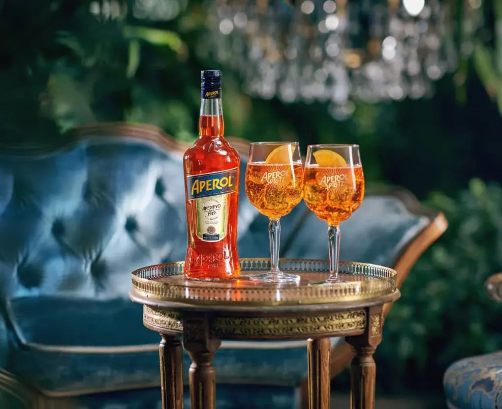 Láhev Aperolu a dvě sklenice Aperol Spritz s plátkem pomeranče na ozdobném stolku, v pozadí modré sametové křeslo a zelené rostliny.