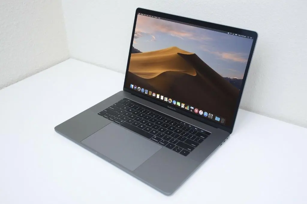 Fotografie zachycuje Apple MacBook Pro umístěný na bílém stole s čistým pozadím. Laptop má tenký a elegantní design typický pro Apple produkty, s velkou obrazovkou zobrazující pracovní plochu operačního systému macOS. Na obrazovce je vidět tapeta s pouštní scenérií, což dodává fotce estetický vzhled. Klávesnice je černá, kontrastující s kovovým tělem notebooku. Celkový vzhled je moderní a minimalistický, což zdůrazňuje prémiovou kvalitu a stylovost tohoto zařízení.