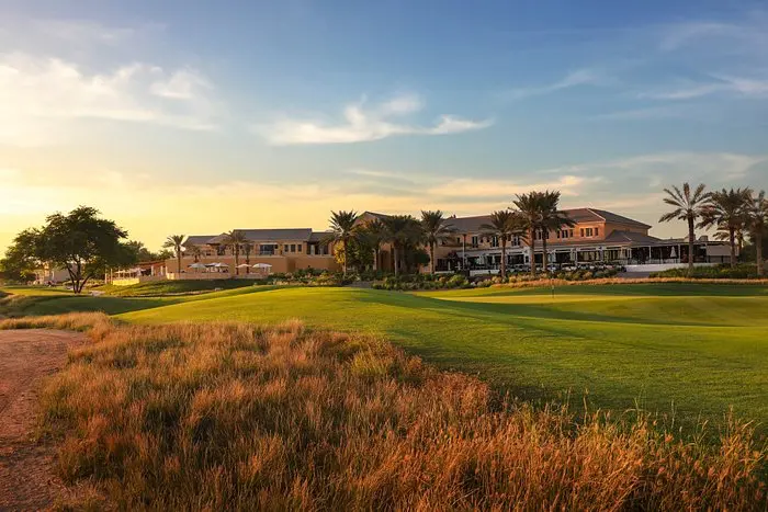 Arabian Ranches Golf Club v Dubaji při západu slunce. Na obrázku je vidět klubovna obklopená palmami a bujnou vegetací. Před klubovnou se rozprostírá zelený trávník golfového hřiště, které přechází do zlatavých travin. Obloha je jemně zabarvena večerním světlem, což vytváří klidnou a příjemnou atmosféru tohoto prestižního golfového klubu.