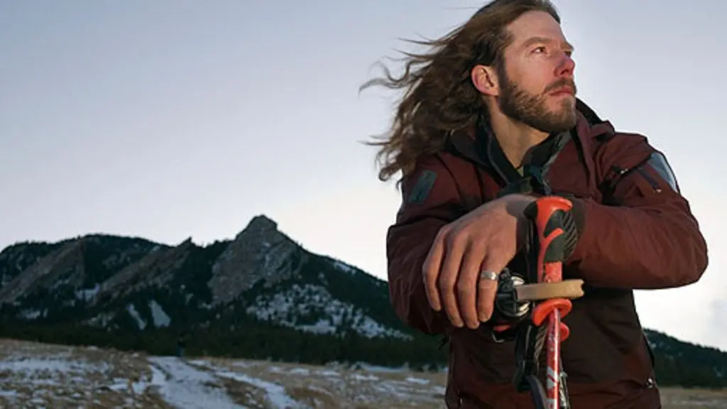 Muž Aron Ralston s dlouhými vlasy a vousy, oblečený v outdoorovém oblečení, drží turistickou hůl a hledí do dálky. V pozadí jsou vidět zasněžené hory a nebe na soumraku.