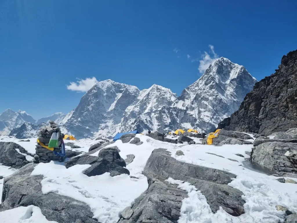 Zasněžený základní tábor s barevnými stany na Mount Everestu, obklopený strmými horskými vrcholy a skalnatým terénem. Jasná modrá obloha doplňuje dramatickou a krásnou horskou scenérii.