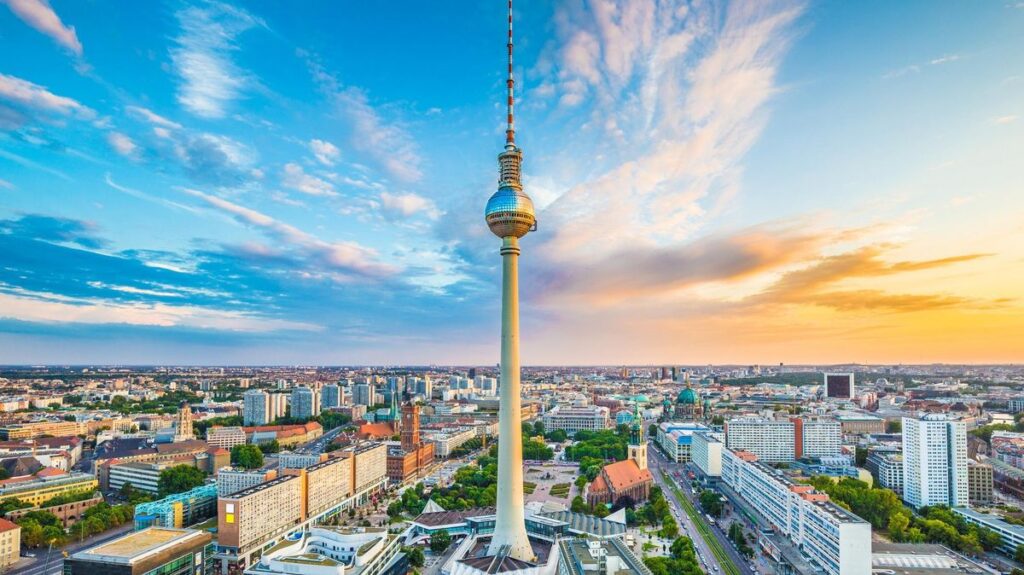 Televizní věž v Berlíně (Berliner Fernsehturm) se tyčí nad městem v pozadí zapadajícího slunce, obklopena moderní městskou zástavbou a historickými budovami. Na obzoru je vidět panorama německé metropole.