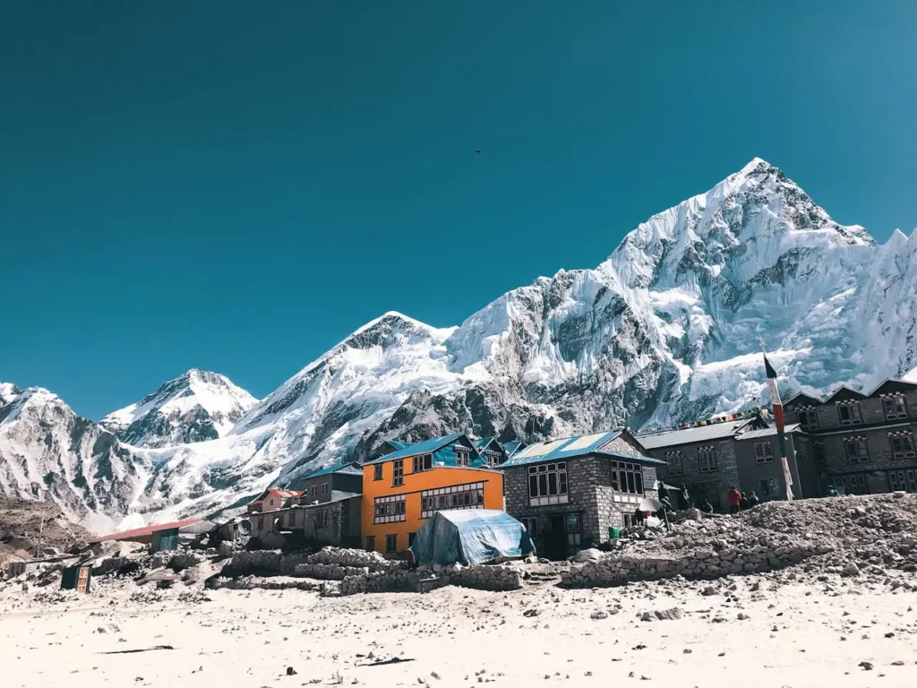 Barevné domy v nepálské vesnici s impozantním pozadím zasněžených vrcholů Himalájí, včetně Mount Everestu. Jasná modrá obloha a horské prostředí vytvářejí krásnou a dramatickou scenérii.