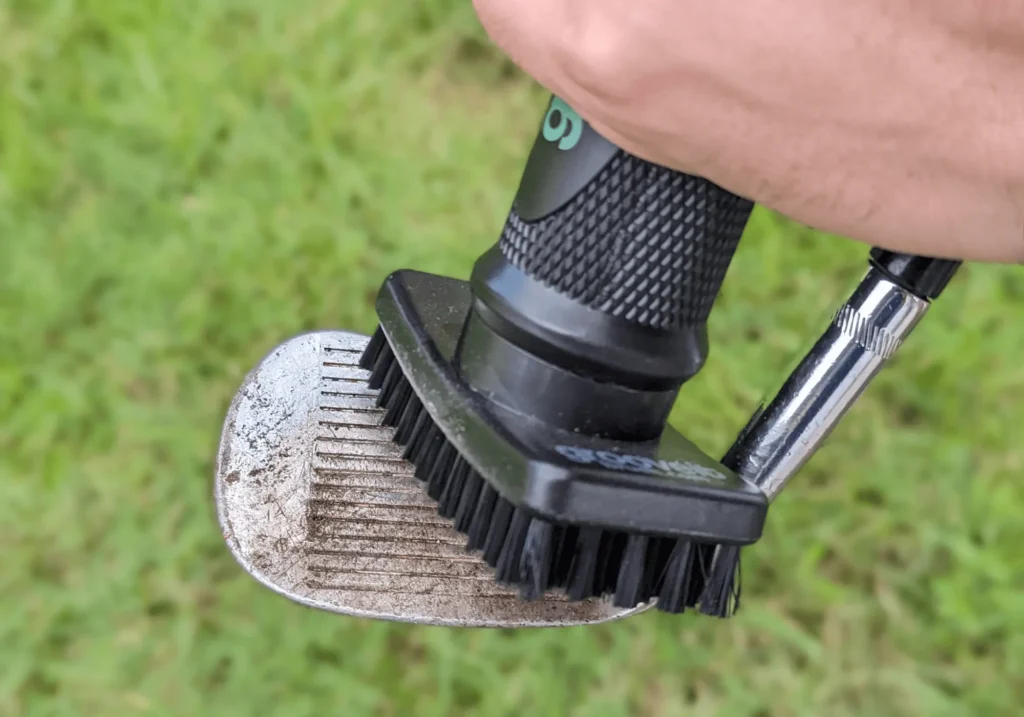 Fotografie zachycuje čištění golfové hole pomocí speciálního kartáče. Na pozadí je vidět tráva, což naznačuje, že se činnost odehrává venku, pravděpodobně na golfovém hřišti. Kartáč má černou rukojeť a je určený k odstranění nečistot z drážek na hlavě golfové hole.