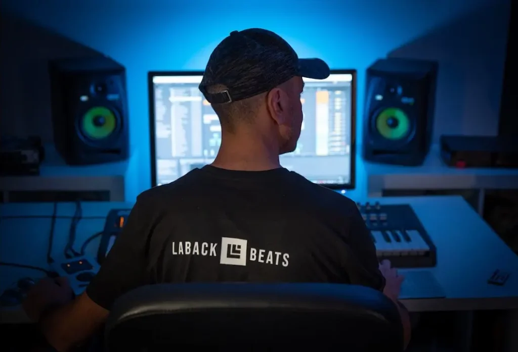 Obrázek zobrazuje osobu sedící před počítačem v nahrávacím studiu. Na zádech má tričko s nápisem "LABACK BEATS". Studio je osvětlováno modrým světlem a na stole před osobou jsou vidět různé zařízení pro hudební produkci, včetně klávesnice a reproduktorů. Vše naznačuje, že se jedná o hudebního producenta či DJ, který pracuje na tvorbě hudby.