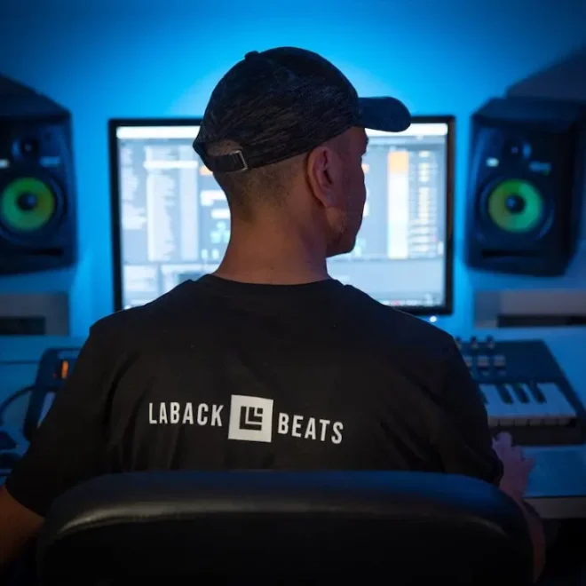 Obrázek zobrazuje osobu sedící před počítačem v nahrávacím studiu. Na zádech má tričko s nápisem "LABACK BEATS". Studio je osvětlováno modrým světlem a na stole před osobou jsou vidět různé zařízení pro hudební produkci, včetně klávesnice a reproduktorů. Vše naznačuje, že se jedná o hudebního producenta či DJ, který pracuje na tvorbě hudby.