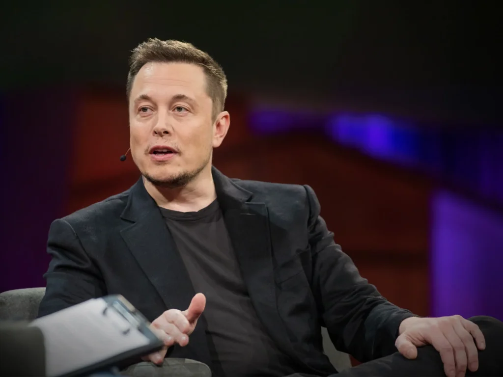 Na obrázku je Elon Musk, zakladatel a CEO společností Tesla a SpaceX, který sedí na pódiu během konference. Má na sobě černé sako a černé tričko a vypadá, že je zapojen do rozhovoru. V pozadí je tmavé, s fialovým osvětlením, což dodává scéně profesionální atmosféru. Elon Musk gestikuluje pravou rukou a působí zaujatě.