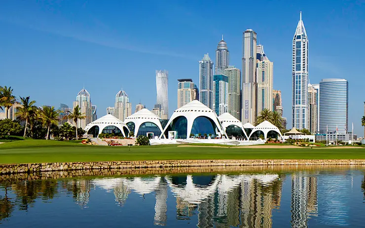 Pohled na Emirates Golf Club v Dubaji, golfové hřiště s moderními budovami v pozadí. V popředí je zelený trávník a vodní plocha, ve které se odráží architektonicky zajímavé stavby včetně vysokých mrakodrapů. Celá scéna je zalitá slunečním světlem, což podtrhuje krásu a modernost města Dubaj.