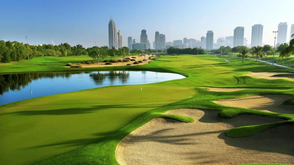 Zelené fairwaye a písečné bunkry na Emirates Golf Club v Dubaji, konkrétně na hřišti Faldo. V pozadí jsou viditelné moderní mrakodrapy dubajského města, zatímco v popředí se nachází vodní překážka a dobře udržovaný trávník. Scéna je prozářená slunečním světlem, zdůrazňující krásu a eleganci tohoto prestižního golfového hřiště.