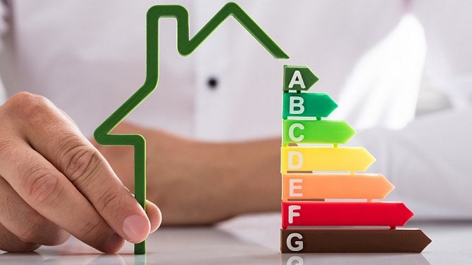 Ruka drží zelenou siluetu domu vedle barevně odstupňovaných šipek znázorňujících energetické třídy budovy od A (nejúspornější) po G (nejméně úspornou).